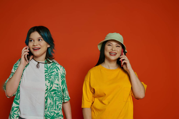 Azjatka i jej nastoletnia córka stojąca obok siebie, pochłonięta oddzielnymi rozmowami telefonicznymi. - Zdjęcie, obraz