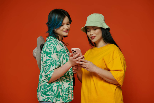Een moeder en haar tienerdochter staan zij aan zij, betoverd door het scherm van een mobiele telefoon in een studio met een oranje achtergrond. - Foto, afbeelding