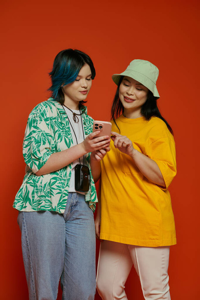 Una madre asiática y su hija adolescente de pie juntas, absortas mientras miran un teléfono celular sobre un fondo naranja. - Foto, Imagen