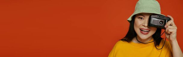 Une femme asiatique tient une caméra sur son visage dans un studio sur fond orange. - Photo, image