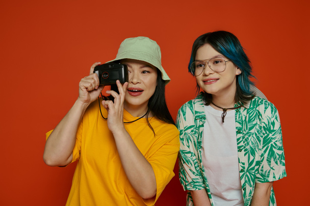 An Asian mother and her teenage daughter stand together in a studio, showcasing unity and familial bond on an orange background. - Photo, Image