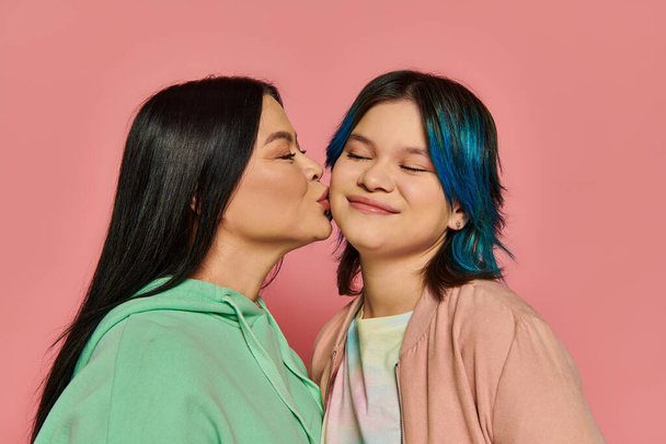 moeder en tiener meisje met levendige blauw haar delen een tedere kus tegen een roze achtergrond. - Foto, afbeelding