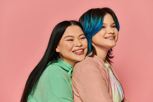 Азиатская мать и дочь-подросток в повседневной одежде, обе с голубыми волосами, радостно обнимающие друг друга в студии на розовом фоне. - Фото, изображение