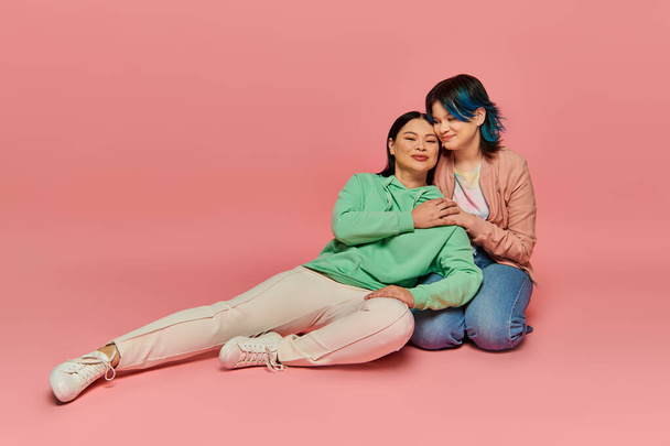 Asian mother and teenage daughter in casual wear sit closely together on a pink background. - Photo, Image