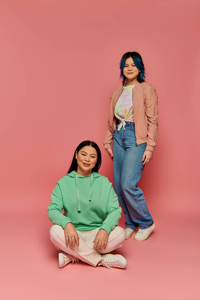 Asian mother and her teenage daughter, dressed casually, standing together against a pink background in a studio setting. - Photo, Image