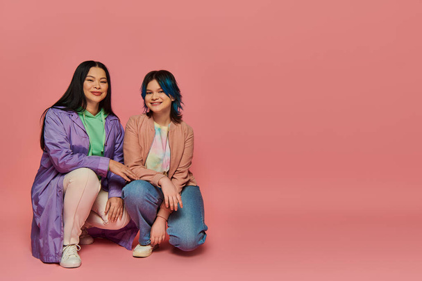 Asian mother and teenage daughter in casual wear sitting closely next to each other on a vibrant pink background. - Photo, Image