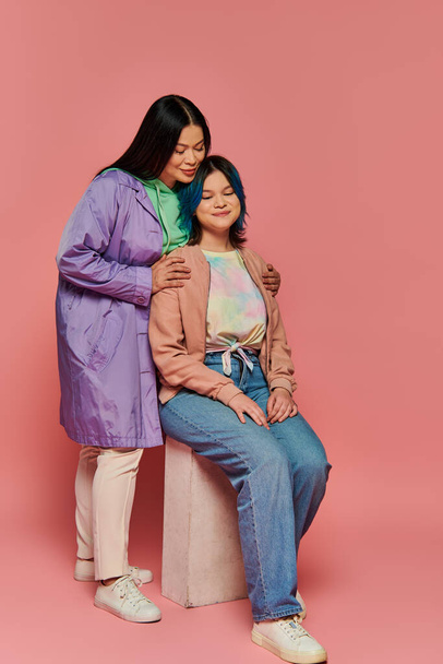 Asyalı bir anne ve genç kızı, her ikisi de gündelik giysiler içinde, canlı pembe bir arka plana karşı bir bankta birlikte oturuyorlar.. - Fotoğraf, Görsel