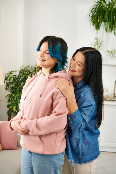 Een Aziatische moeder en haar tienerdochter staan zij aan zij in een gezellige woonkamer en delen een moment van verbondenheid en warmte. - Foto, afbeelding