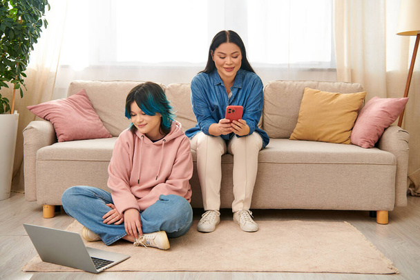 Asyalı anne ve genç kızı, günlük giysiler içinde, bir kanepede oturuyorlar ve bir cep telefonu ekranına odaklanıyorlar.. - Fotoğraf, Görsel