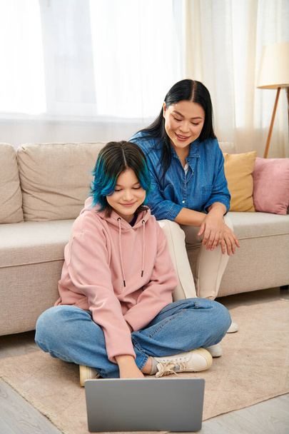 Eine asiatische Mutter sitzt auf dem Boden neben ihrer Tochter im Teenageralter, die auf einen Laptop-Bildschirm fixiert ist und sich an der Technik festhält. - Foto, Bild