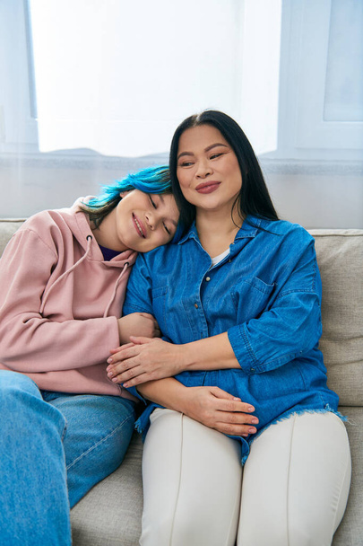Μια Ασιάτισσα μητέρα και η έφηβη κόρη της με καθημερινά ρούχα, κάθονται πάνω σε έναν καναπέ, μοιράζονται ποιοτικό χρόνο μαζί στο σπίτι.. - Φωτογραφία, εικόνα