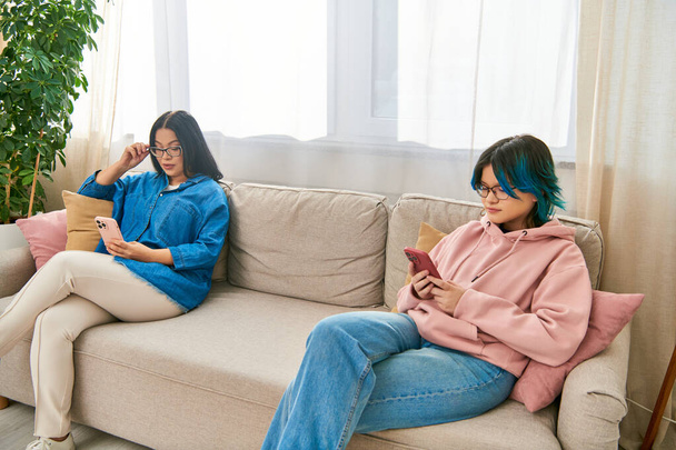 Ασιάτισσα μητέρα και έφηβη κόρη με καθημερινά ρούχα κάθονται σε έναν καναπέ, ασχολούνται με τα τηλέφωνά τους, περνώντας χρόνο μαζί στο σπίτι. - Φωτογραφία, εικόνα