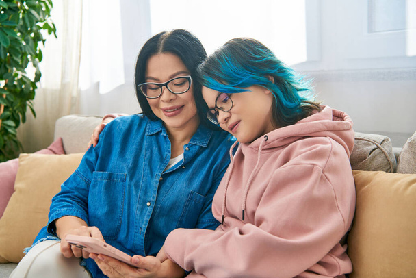 Asiatische Mutter und Tochter sitzen in lässiger Kleidung auf einer Couch, konzentrieren sich auf einen Handybildschirm und verbringen Zeit miteinander.. - Foto, Bild