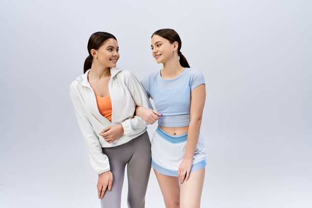 Zwei hübsche, brünette junge Frauen in sportlicher Kleidung stehen nebeneinander und strahlen auf grauem Studiohintergrund Zuversicht und Freundschaft aus.. - Foto, Bild