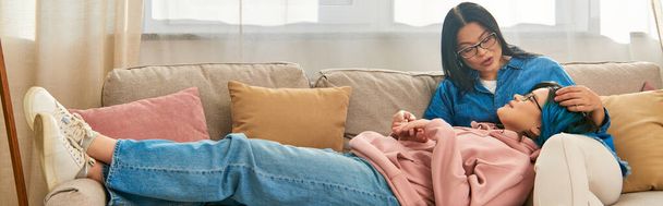 Μια Ασιάτισσα μητέρα και η έφηβη κόρη της κάθονται σε έναν καναπέ, απολαμβάνοντας μια ζεστή στιγμή στο σπίτι. - Φωτογραφία, εικόνα
