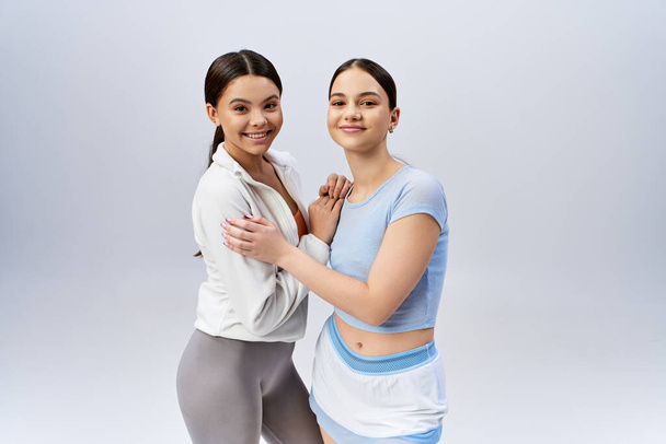 Zwei hübsche, sportliche Teenager-Mädchen, eine brünett, posieren selbstbewusst zusammen in einem Studio vor grauem Hintergrund. - Foto, Bild