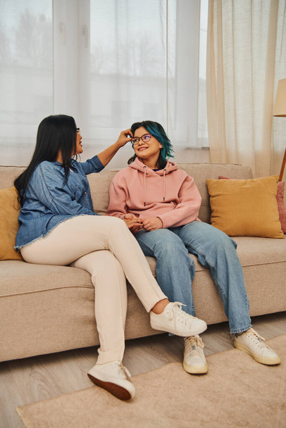 Asiatische Mutter und Tochter in lässiger Kleidung unterhalten sich lebhaft, während sie zu Hause auf einer gemütlichen Couch sitzen. - Foto, Bild
