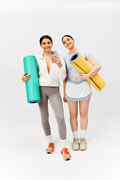 İki güzel genç kız, bir esmer, spor kıyafetleriyle birlikte gri stüdyo arka planında yoga minderleri tutuyorlar.. - Fotoğraf, Görsel