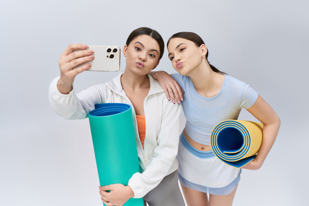 Dwóch ładnych nastoletnich przyjaciół, sportowa brunetka i sportowy strój, uwieczniają selfie z matą do jogi w studio. - Zdjęcie, obraz