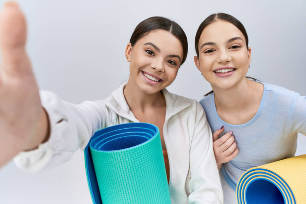 Two pretty and brunette teenage girls, female friends, in sportive attire standing next to each other with yoga mats in a studio on a grey background. - Photo, Image