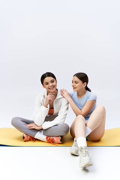 Two pretty, brunette teenage girls in sportive attire sitting on a mat, holding hands in unity and friendship. - Photo, Image