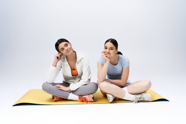 Две симпатичные брюнетки-девочки в спортивном костюме сидят на коврике для йоги, разделяя момент расслабления и общения.. - Фото, изображение