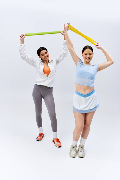 Zwei hübsche brünette Teenager-Mädchen in sportlicher Kleidung stehen zusammen, verströmen Stärke und Freundschaft auf grauem Studiohintergrund. - Foto, Bild