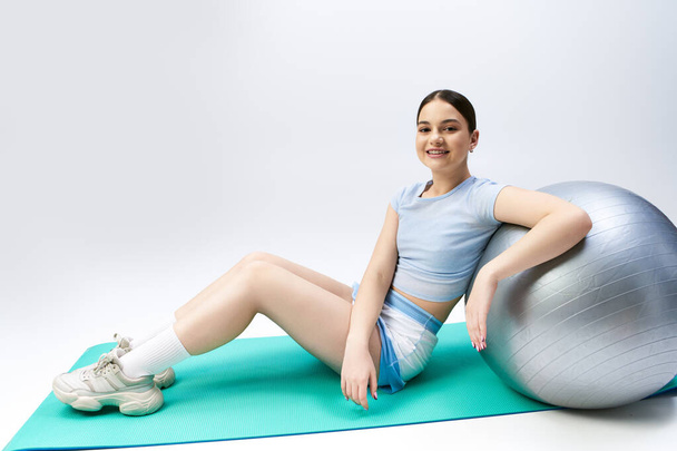 Ένα όμορφο, μελαχρινό κορίτσι με σπορ ενδυμασία κάθεται με χάρη πάνω από μια μπάλα γυμναστηρίου σε ένα σκηνικό στούντιο - Φωτογραφία, εικόνα
