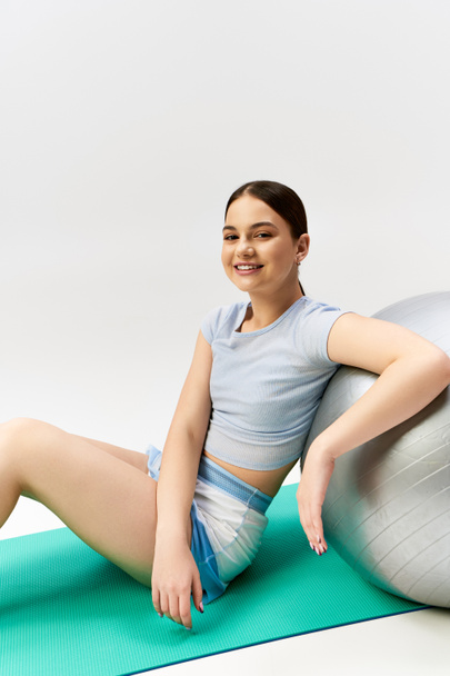 Una guapa adolescente morena vestida con un atuendo deportivo equilibra graciosamente una pelota de yoga en un estudio - Foto, imagen