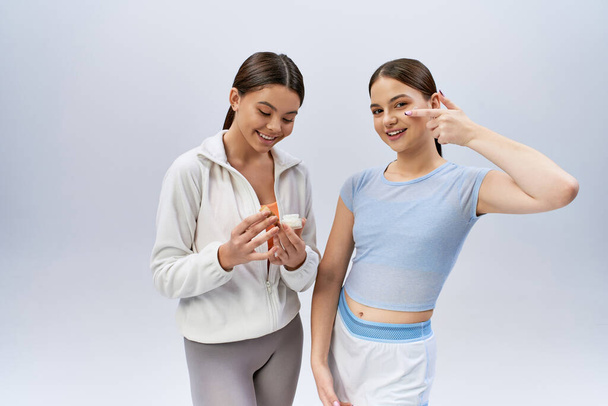 Zwei hübsche, brünette Teenager-Mädchen in sportlicher Kleidung stehen in selbstbewusster Pose nebeneinander auf grauem Studiohintergrund. - Foto, Bild