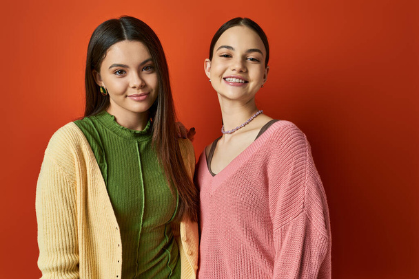 Two pretty and brunette teenage girls in casual attire standing next to each other in front of a vibrant red wall. - Photo, Image