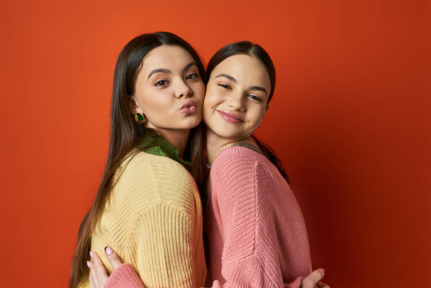 Δύο όμορφες, μελαχρινές έφηβες κοπέλες με απλή ενδυμασία αγκαλιάζονται τρυφερά μπροστά σε έναν ζωντανό κόκκινο τοίχο.. - Φωτογραφία, εικόνα