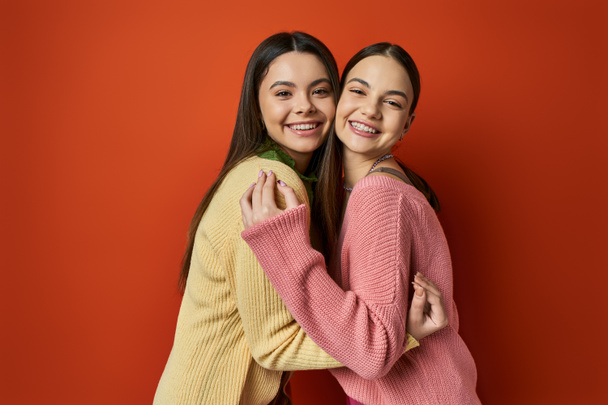 Kaksi nättiä, ruskeaverikkö teini tytöt rento puku halaavat toisiaan edessä elinvoimainen punainen seinä. - Valokuva, kuva