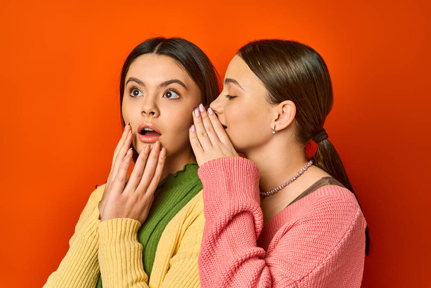 Két csinos, barna tinédzser lány alkalmi ruhában egymás mellett áll nyitott szájjal meglepve egy narancssárga stúdió hátterében.. - Fotó, kép