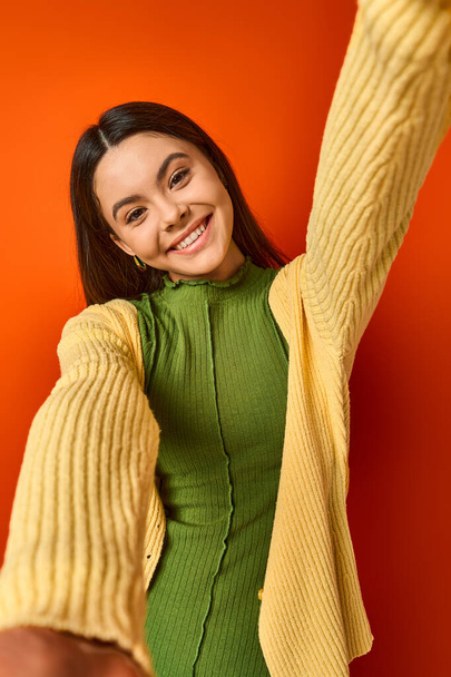 A brunette teenage girl wearing a green dress and a yellow cardigan poses elegantly on an orange background in a studio. - Photo, Image