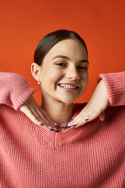 ピンクのセーターのブルネットの十代の少女はスタジオの設定でオレンジ色の背景に対して明るく微笑んでいます. - 写真・画像