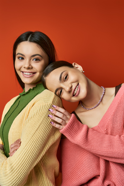 Two pretty, brunette teenage girls wearing casual attire hugging each other in front of a vivid red wall in a studio. - Photo, Image