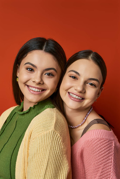 Δύο όμορφα, μελαχρινά έφηβα κορίτσια στέκονται πλάι-πλάι μπροστά σε ένα ζωντανό κόκκινο τοίχο, αποπνέοντας στυλ και φιλία. - Φωτογραφία, εικόνα