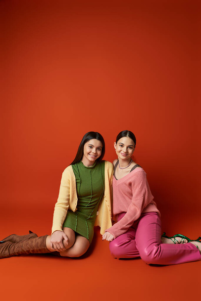 Twee mooie, brunette tienermeisjes zitten op de vloer in een studio, sierlijk poserend voor een foto tegen een oranje achtergrond. - Foto, afbeelding