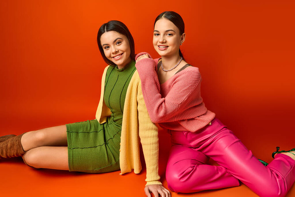 Dvě hezké, brunetky dospívající dívky sedí na zemi v neformálním oblečení a zaujímají pózu pro fotku na oranžovém pozadí. - Fotografie, Obrázek