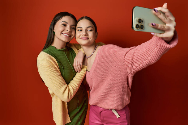 Two pretty teenage friends capturing a moment with a cell phone in a studio against an orange background. - Photo, Image