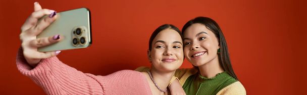 Zwei junge Frauen, Freundinnen, machen ein Selfie mit einem Smartphone vor orangefarbenem Hintergrund in einem Studio-Setting. - Foto, Bild