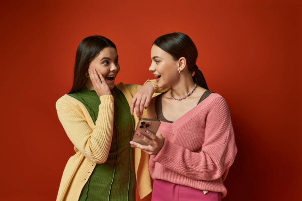 Twee mooie brunette tienermeisjes staan dichtbij, verdiept in een mobiele telefoon scherm, het delen van een moment van verbinding. - Foto, afbeelding