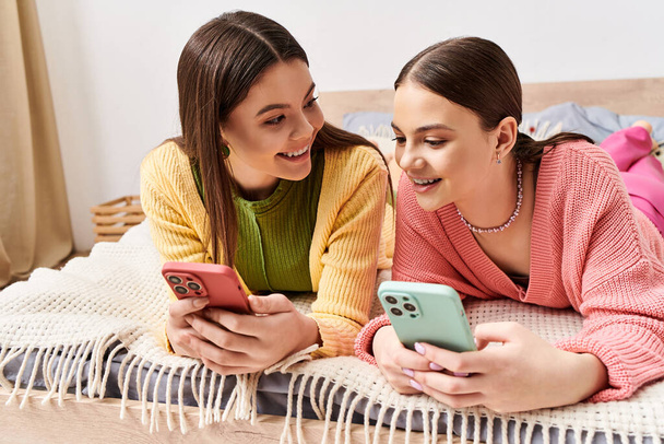 Deux jeunes femmes en tenue décontractée se prélassant sur un lit, absorbées dans leurs téléphones cellulaires. - Photo, image