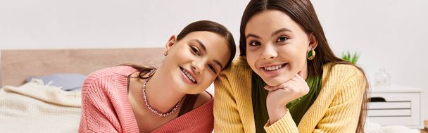 Dvě hezké dospívající dívky v neformálním oblečení sedí těsně vedle sebe na posteli doma a užívají si společnost. - Fotografie, Obrázek