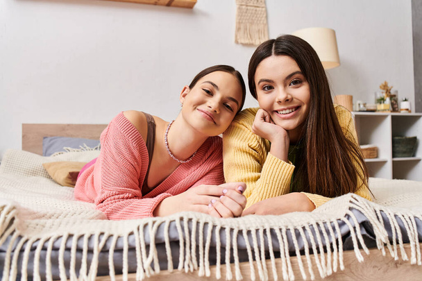 Twee vriendinnen, mooie tienermeisjes in casual kleding, liggend op een bed, glimlachend naar de camera in een sereen moment. - Foto, afbeelding