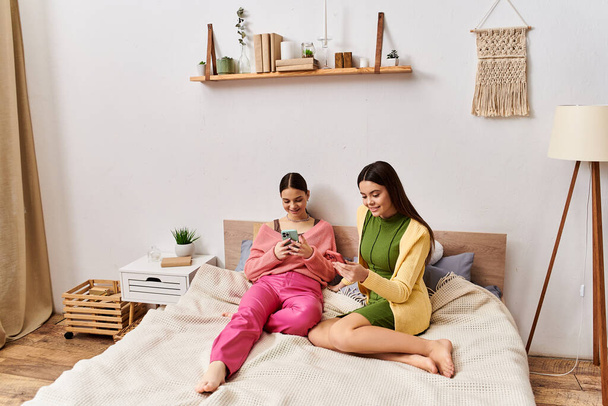 Deux jeunes femmes en tenue décontractée assises sur un lit, englouties dans un téléphone portable. - Photo, image