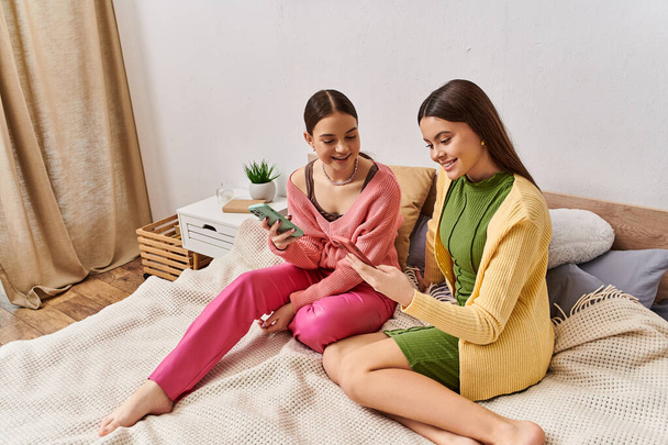 Két tinédzser lány ül az ágyon, lefoglalva egy mobiltelefonnal.. - Fotó, kép