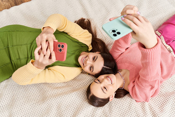 Δύο νεαρές γυναίκες με casual ρούχα απολαμβάνουν μια στιγμή χαλάρωσης καθώς ξαπλώνουν δίπλα δίπλα σε ένα άνετο κρεβάτι. - Φωτογραφία, εικόνα