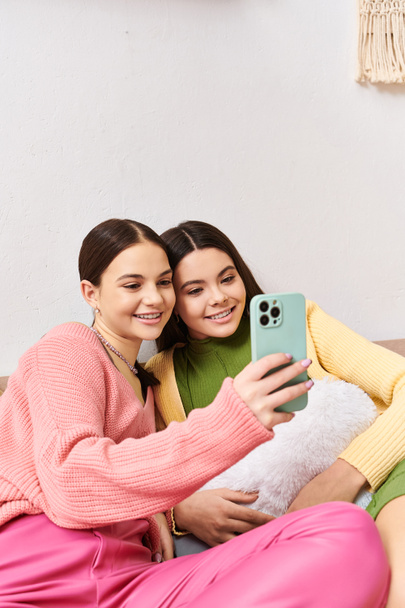 Zwei hübsche Teenager-Mädchen in lässiger Kleidung sitzen auf einer Couch und fangen einen lustigen Moment ein, indem sie zusammen ein Selfie machen. - Foto, Bild